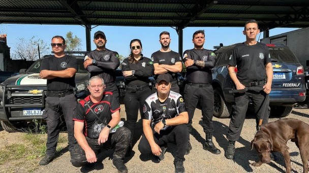 Na foto, sete homens com roupas pretas e uma mulher também vestida com a farda preta posam para a foto. No canto direito um cachorro labrador está junto aos policiais penais.