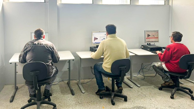 Na foto, três homens estão sentados na frente de computadores
