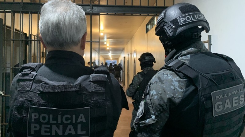 Na foto, um homem com cabelos brancos esta ao lado de um outro homem com vestimentas da Polícia Penal